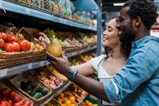 happy asian woman looking at fruits near cheerful african american man in store
